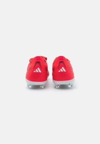 Para jasnoczerwonych korków piłkarskich z białymi logo Adidas widocznymi na piętach, zwróconych plecami, na jednolitym białym tle.