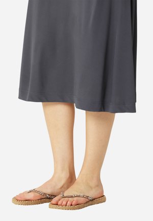 Lower legs wearing a dark skirt and beige braided flat sandals with beaded straps on a white background.