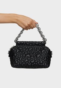 Mano che tiene una borsa nera testurizzata con forme arrotondate e un'impugnatura a catena spessa in argento su uno sfondo semplice.