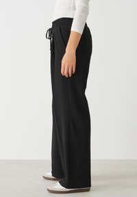 Zwarte wijde broek met een elastische tailleband en trekkoord. Glad materiaal, relaxte pasvorm, gecombineerd met witte sneakers met een bruine zool.