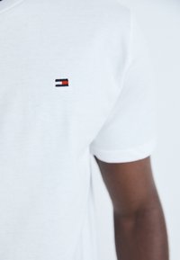 Primer plano de una persona que lleva una camiseta blanca con un pequeño logo rectangular en azul marino, blanco y rojo en el pecho izquierdo.