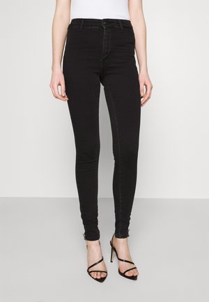 Femme portant un jean skinny noir taille haute et des sandales noires à talons hauts et brides, sur un fond blanc uni.