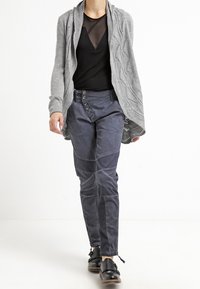 Grå stickad kofta med flätmönster och öppen front, lager på en svart genomskinlig topp. Mörkblå avsmalnande byxor med knappdetaljer och kontrasterande paneler.