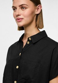 Črna majica iz teksturirane tkanine, z ovratnikom, kratkimi rokavi in zlatimi gumbi. Dva žepa spredaj.