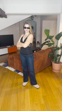 Czarny top bez rękawów, niebieskie rozszerzane dżinsy i beżowe obuwie. Wnętrze z brązową sofą, doniczkową rośliną i drewnianą podłogą.