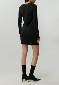 Schwarzes Langarmkleid mit tailliertem Schnitt, knielang, aus einem strukturierten Stoff. Kombiniert mit schwarzen Knöchelsocken und Absatzschuhen.