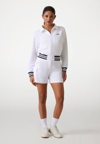 Witte zip-up jack met een kraag en marineblauwe gestreepte accenten, gecombineerd met witte shorts met zakken en een vergelijkbaar ontwerp van de tailleband.