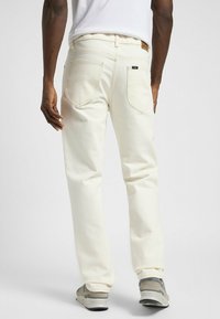 Jeans en coton blanc avec une coupe droite, comportant des poches arrière et une étiquette de marque en cuir marron au niveau de la taille. Des baskets grises sont portées.