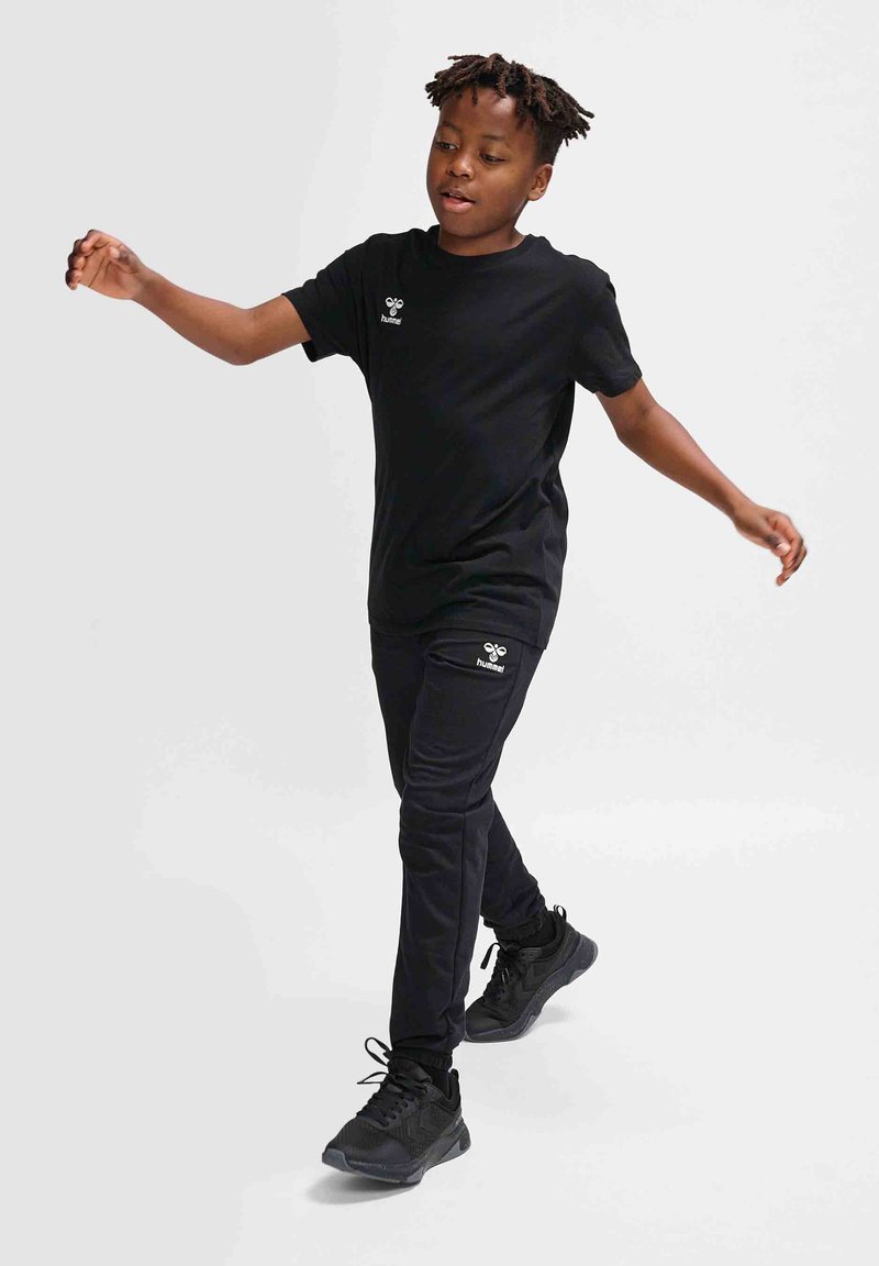 Schwarzes T-Shirt mit kurzen Ärmeln und schwarze Jogginghose, die ein kleines Logo auf der Brust und am Bein aufweist. Das Modell trägt schwarze Laufschuhe.