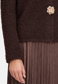 Brauner strukturierter Pullover mit rundem Halsausschnitt und langen Ärmeln, kombiniert mit einem gefalteten Rock aus braunem Wildleder. Eine dekorative Stoffblume als Brosche schmückt den Pullover.