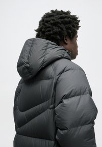 Czarna kurtka puchowa z kapturem, z pikowanymi panelami i gładką, błyszczącą teksturą. Widok z tyłu ukazuje luźny krój i subtelne detale.