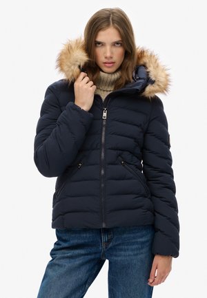 Marineblauwe gewatteerde jas met een met bont afgewerkte capuchon, ritszakken en een doorgestikte uitstraling, gedragen over een crèmekleurige trui en blauwe jeans.