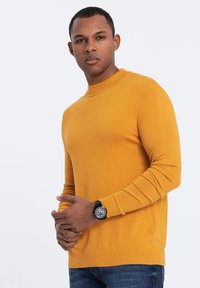 Mustard yellow sweater with a round neckline, ribbed cuffs, and a smooth texture. Worn with dark denim jeans and a black watch.
