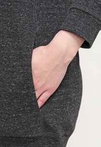 Dark gray sweatshirt fabric featuring a side pocket with a textured, heathered pattern. Cuffs are fitted and trimmed with neat stitching.