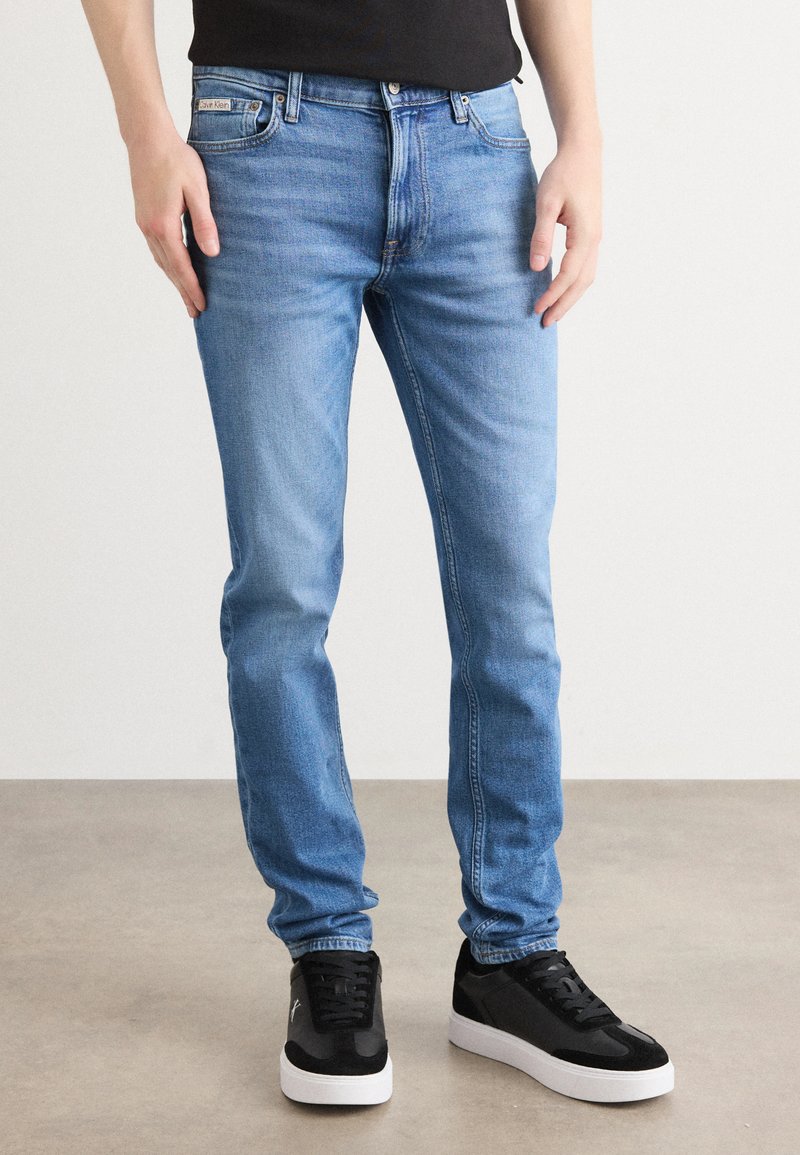 Jean en denim bleu clair avec une coupe slim, dotés de poches avant et d'une fermeture éclair. Associés à des baskets noires avec des semelles blanches.