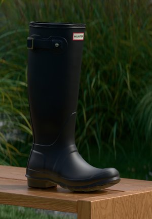 Black rubber boot with a round toe and textured sole. Features a buckle detail and brand label on the upper side. Smooth surface and tall design.
