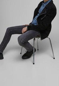 Černá semišová bunda přes džínsovou košili, šedé slim-fit džíny a černé boty. Sedí na židli s chromovými nohami na neutrálním pozadí.