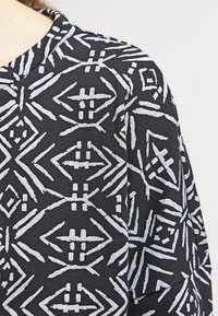Camisa de tela negra con un patrón geométrico blanco que incluye diversas formas, como diamantes y flechas, creando una apariencia texturizada.