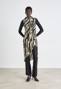 Robe asymétrique à imprimé abstrait jaune et noir, associée à un pantalon noir ajusté et des talons noirs pointus. Présente des détails froncés.