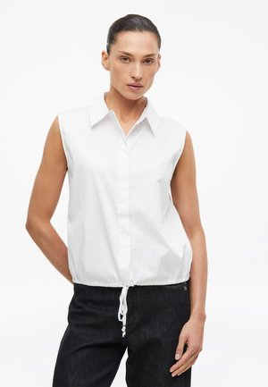 Vrouw met donker haar in een witte mouwloze top met kraag en aantrekkoord in de taille en zwarte broek tegen een effen witte achtergrond.