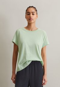 Hellgrünes, kurzärmeliges T-Shirt mit lockerem Schnitt und Rundhalsausschnitt; weiche, glatte Textur; kombiniert mit dunklen, lockeren Hosen.