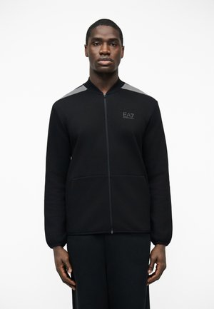 Férfi fekete cipzáras dzsekiben, szürke vállrészletekkel és az "EA7 Emporio Armani" logóval a mellkasán, egyszerű fehér háttér előtt állva.