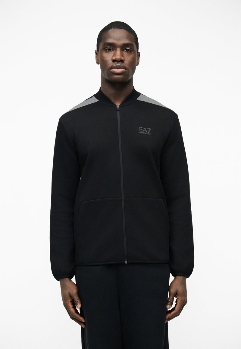 Άνδρας που φοράει μαύρο μπουφάν με φερμουάρ και γκρι λεπτομέρειες στους ώμους, με το λογότυπο "EA7 Emporio Armani" στο στήθος, στέκεται μπροστά σε απλό λευκό φόντο.