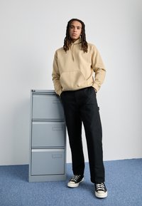 Beige hoodie met een buidelzak, zwarte loszittende broek en zwarte canvas sneakers met witte zolen. Staande naast een grijze archiefkast.