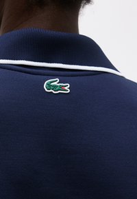 Άτομο που φοράει μπλε σκούρο μπλουζάκι πόλο με λευκή διακόσμηση και ένα μικρό πράσινο λογότυπο κροκόδειλου στο πάνω μέρος της πλάτης κοντά στον γιακά.