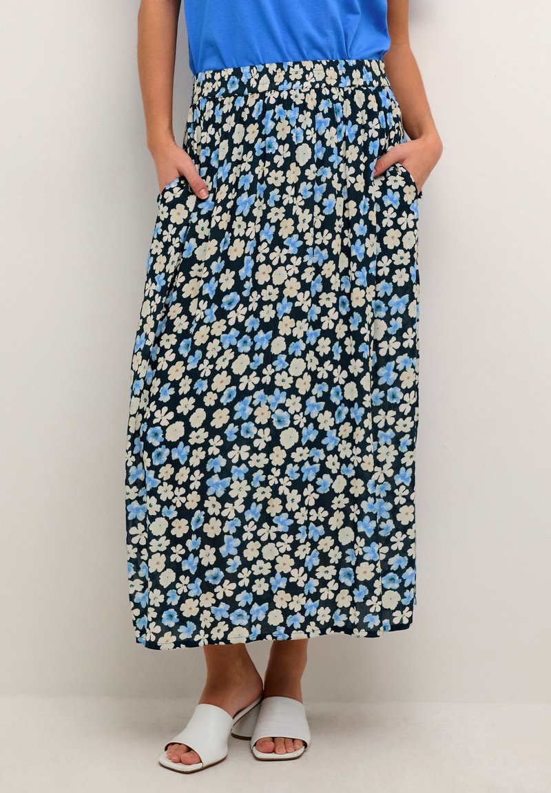 Bloemenmaxirok in marineblauw met blauwe en crèmekleurige bloemen. Heeft een elastische tailleband en zijzakken. Draagt witte instapsandalen.