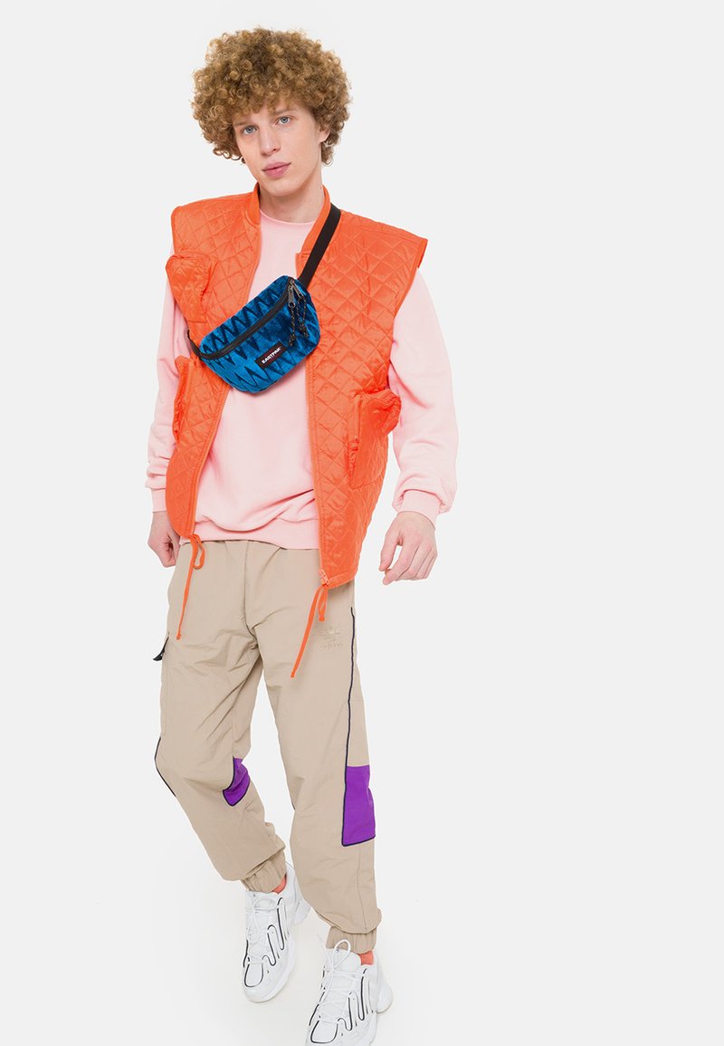 Gilet arancione trapuntato sopra una felpa rosa, pantaloni beige con accenti viola e una borsa a tracolla blu con motivo. Sneakers bianche completano il look.