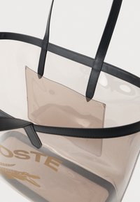 Prozirna PVC torba s crnim kožnim trakama i akcentima. Sadrži zlatni logo "LACOSTE" i malu unutarnju džepu.