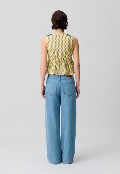 Top giallo senza maniche a peplo con pois blu navy e vita arricciata, abbinato a jeans a gamba larga di un blu chiaro, con tasche posteriori visibili.