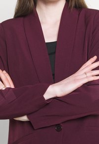 Marynarka burgundowa o luźnym fasonie, z kołnierzem z klapą, zapięciem na pojedynczy guzik i długimi rękawami. Teksturowana tkanina, płaskie kieszenie z przodu.