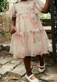 Robe rose clair avec des motifs floraux, manches volantées et jupe à volants. Associée à des chaussures Mary Jane blanches. Photographie prise sur des marches en pierre.