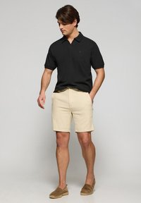 Camiseta polo negra con cuello corto, pantalones cortos color crema y alpargatas marrones. Textura suave y corte casual. Diseño simple sin patrones.