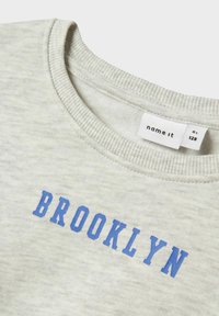 Ljust grå sweatshirt med ribbat krage och blå "BROOKLYN" text tryckt över bröstet, visar inarbetad varumärkesetikett inuti kragen.