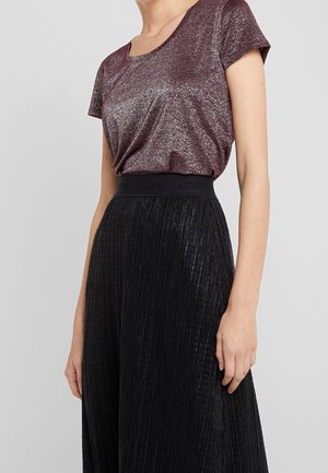 Femme portant un haut violet scintillant à manches courtes, rentré dans une jupe noire plissée taille haute avec une large ceinture.