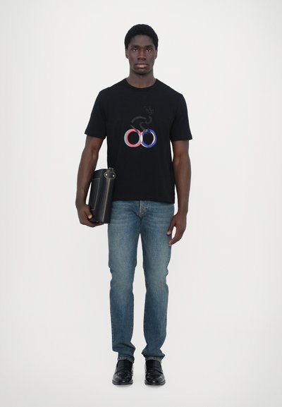 T-shirt noir avec un design de cercles olympiques multicolores. Porté avec un jean bleu et des chaussures noires, tenant un sac rectangulaire noir.