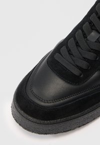 Gros plan sur l'avant d'une basket noire avec des textures en daim et en cuir, des lacets arrondis et une semelle en caoutchouc épaisse et texturée, sur fond blanc.