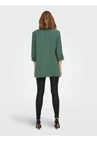 Vihreä tunika, jossa on käärityt hihat ja suora leikkaus. Yhdistettynä mustiin legginsseihin ja korkokenkiin. Tasainen, kevyt kangas.