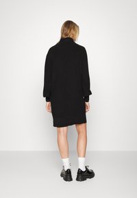 Svart oversize stickad klänning med hög krage och långa ärmar. Slät textur, fåll ovanför mitten av låret, tillsammans med kraftiga svarta skor.