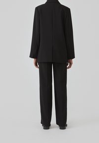 Personne debout de dos, portant un blazer noir et un pantalon noir assorti sur un fond clair uni.