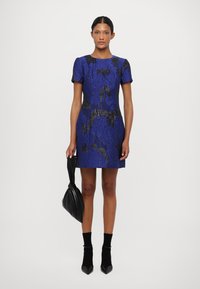 Zila mežģīņu kleita ar īsām piedurknēm un melnu ziedu rakstu, kombinēta ar melnu somu. Tai ir pieguļoša forma un ceļa garuma griezums.