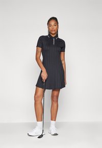 Mustagolfi kleit lühikeste varrukatega, ribakujuline tekstuur ja polo krae valge accentsiga. Mudel hoiab golfikeppi ja kannab valgeid ujumissusse.