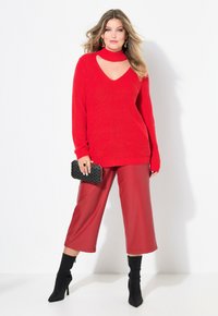 Pull en tricot rouge avec un décolleté en découpe, associé à un pantalon bordeaux taille haute et à jambes larges. Le mannequin tient une pochette texturée noire. Talons noirs.