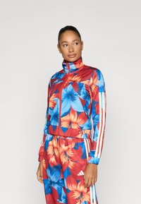 Φόρμα με φλοράλ μοτίβο σε κόκκινο, μπλε και πορτοκαλί. Διαθέτει ψηλό γιακά, φερμουάρ και λευκές λωρίδες στα πλάγια στα μανίκια και το παντελόνι.