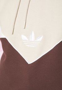 Hoodie med en beige övre del, brun nedre del, vit kant och broderad logotyp. Materialet är mjukt med en strukturerad finish.