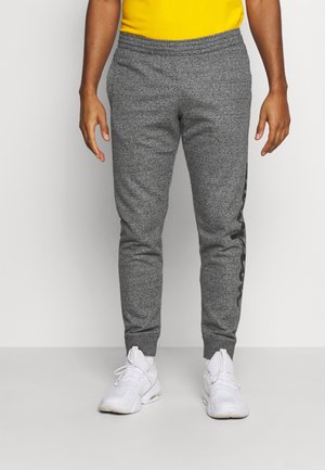 Persona con pantalones deportivos grises con texto negro lateral, zapatillas blancas y una camiseta amarilla, de pie contra un fondo claro y sencillo.