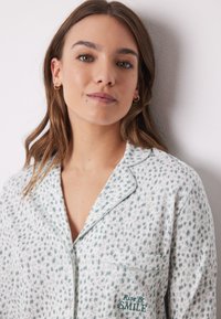 Vit pyjamasöverdel med grönt mönster och broderi "Rise & SMILE". Har en klassisk krage, ficka och piping längs kanterna.
